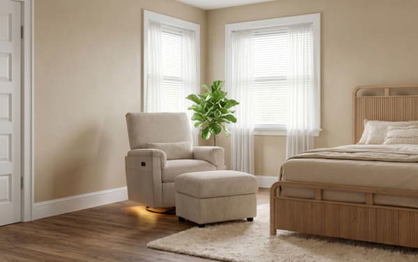 Beige upholstered recliner chair with matching ottoman, wooden frame bed with neutral bedding, hardwood floor, and natural light from windows in a cozy bedroom.